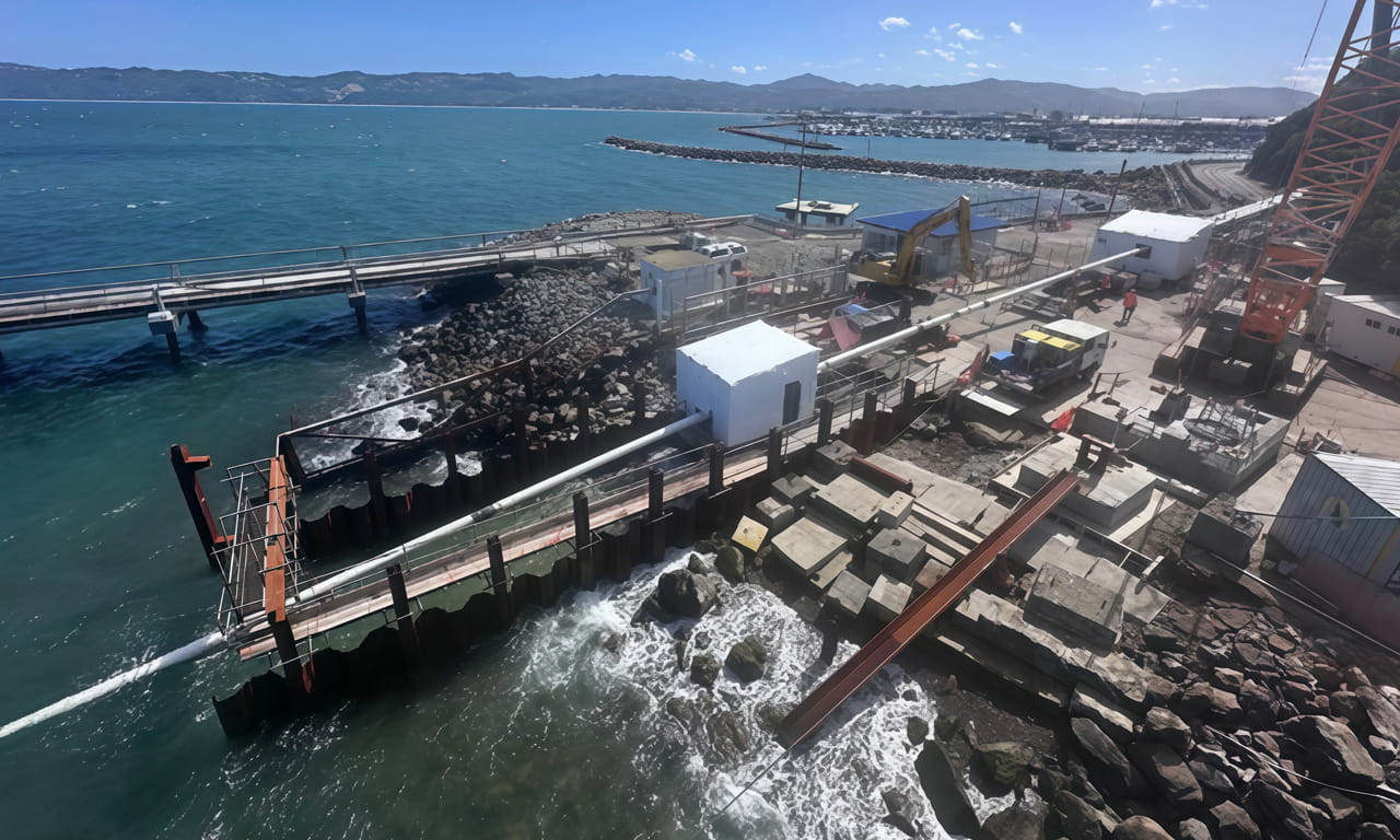 Ongoing coastal construction including cranes and excavators, alongside marine piling works.